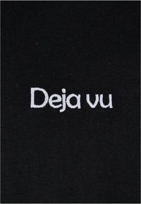 Juodas audinys su baltu siuvinėtu tekstu, kuris sako "Deja vu", centre ir su švelnia tekstūra. Paprastas ir minimalistinis dizainas.
