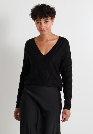 Un pull en maille noir avec un décolleté en V profond, un motif à rayures verticales et des manches côtelées, associé à une jupe noire lisse.