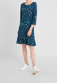 Blå klänning med blommönster, 3/4 ärmar och utsvängd nederkant. Tillverkad av mjukt material, bärs med vita sneakers på en neutral bakgrund.