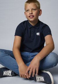Mörkblå pikétröja med korta ärmar, tvåknappsslutning och en liten logotyp; bärs med ljusblå jeans och mörkblå sneakers.