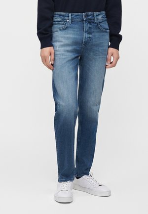 Personne portant un jean bleu délavé à jambes droites, des baskets blanches et un haut à manches longues foncé, debout devant un fond clair uni.