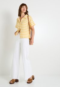 Camisa de manga corta a rayas amarillas y blancas, pantalones anchos blancos, sandalias de plataforma marrones y un bolso pequeño.