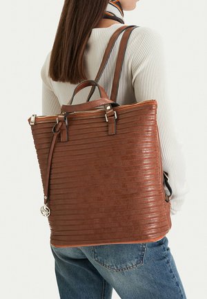 Donna che indossa un maglione crema a coste e jeans blu, porta uno zaino grande in pelle marrone testurizzata con cerniera e manici superiori.