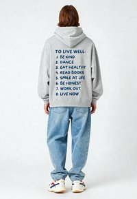 Felpa grigia con cappuccio caratterizzata da testo blu che elenca otto principi per il benessere sul retro. Vestibilità oversize, abbinata a jeans azzurri e sneakers.