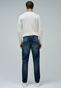 Jeans de mezclilla azul oscuro con corte recto, con un sutil desgastado y dos bolsillos traseros. Combinados con un suéter de punto blanco.