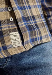 Brązowa i niebieska koszula w kratę z białymi guzikami, z tkanym metkiem z napisem "CROSS JEANS", obok niebieskiego denimu.
