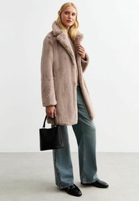 Cappotto in pelliccia sintetica di un beige chiaro con colletto ampio, abbinato a jeans blu a vestibilità comoda e mocassini neri, mentre tiene una piccola borsa nera.