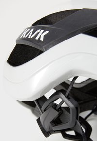 Nahaufnahme eines weißen Kask-Radhelms, der das Logo, die Belüftungsöffnungen und den verstellbaren Kinnriemenmechanismus zeigt.