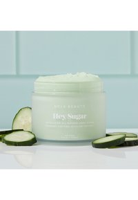 Lys grønn krukke med kroppsskrubb med teksturert lokk. Etiketten leser "Hey, Sugar" og viser agurker på en reflekterende overflate.