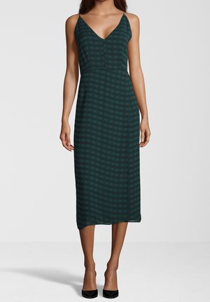 Kobieta w ciemnozielonej, bezrękawnikowej sukience midi z delikatnym kratkowanym wzorem i czarnych szpilkach z wąskim czubkiem, stojąca na neutralnym tle.