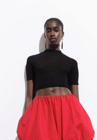 Un top scurt ribbed negru asociat cu o fustă roșie voluminous. Modelul poartă cercei circulari din aur. Fundalul este alb simplu.