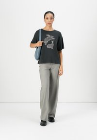 A woman stands against a plain white background, wearing a black shirt featuring rabbit designs, gray wide-leg pants, and holding a blue bag.