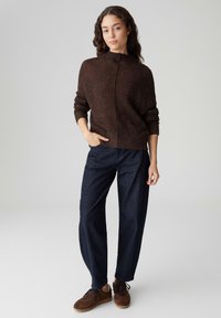 Brauner gestrickter Pullover mit hohem Kragen, kombiniert mit dunkelblauen Jeans mit weitem Bein und braunen Turnschuhen. Die Textur des Pullovers ist weich und gemütlich.