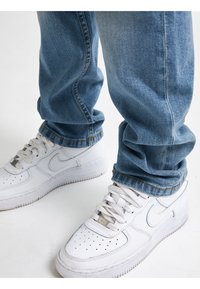 Zapatillas Nike Air Force 1 de cuero blanco con puntera perforada y suela de goma gruesa. Llevadas con jeans de mezclilla azul claro, de corte ligeramente holgado.