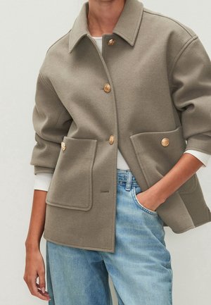 Person wearing taupe wool jacket with gold buttons, large front pockets, white long-sleeve shirt, and light blue jeans.