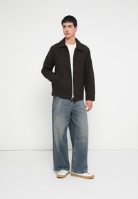 Braune Jacke aus Wollmischung mit Kragen, einer Brusttasche und langen Ärmeln, kombiniert mit hellblauen Jeans mit weitem Bein und weißen Turnschuhen.