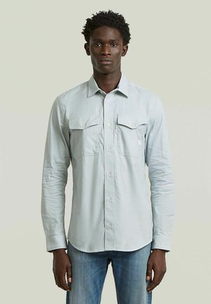 Uomo in piedi che indossa una camicia grigia chiara con bottoni e due tasche sul petto, abbinata a jeans blu, su uno sfondo semplice.