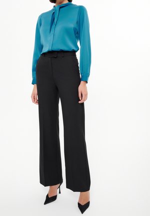 Vrouw draagt een groenblauwe blouse met lange mouwen, zwarte wijde broek met hoge taille en zwarte puntige hakken, staand op een witte achtergrond.