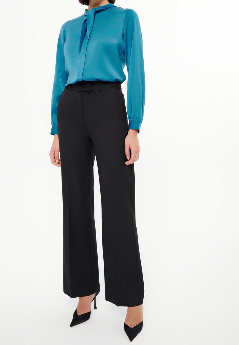 Femme portant un chemisier turquoise à manches longues, un pantalon noir taille haute et jambes larges, ainsi que des talons noirs à bouts pointus, debout sur un fond blanc.
