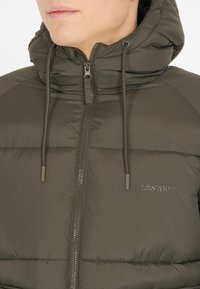 Polstret olivengrøn jakke med hætte, der har en frontlynlås, justerbare snore og logo på brystet. Glat tekstur.