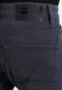 Mörka denimjeans med en texturerad svart läderetikett som har "RAW." Synlig bakficka med böjd sömdetalj.