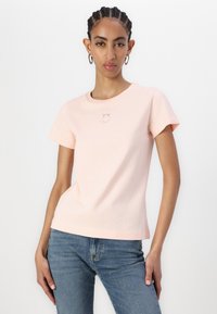 Rožinė medvilninė t-shirt su trumpomis rankovėmis, apvaliu iškirpimu ir mažo logotipo siuvinėjimu priekyje. Dėvima su mėlynomis džinsomis.