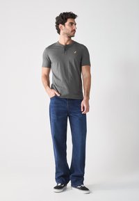 Grijze korte mouw henley-shirt met drie knopen en een klein logo, gecombineerd met losse donkerblauwe jeans en zwarte sneakers.