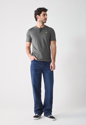 Grijze korte mouw henley-shirt met drie knopen en een klein logo, gecombineerd met losse donkerblauwe jeans en zwarte sneakers.