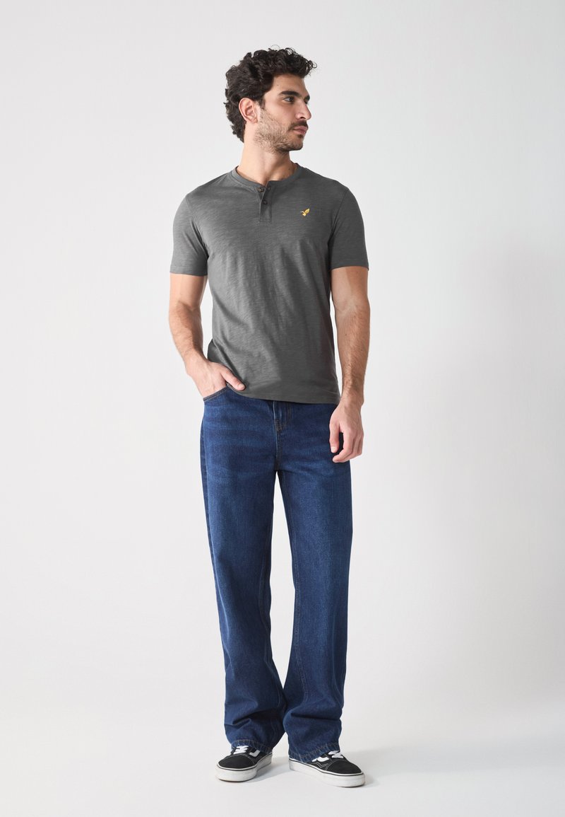 Grijze korte mouw henley-shirt met drie knopen en een klein logo, gecombineerd met losse donkerblauwe jeans en zwarte sneakers.