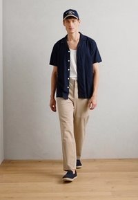 Camicia azzurra a maniche corte sopra una canotta bianca, pantaloni beige e scarpe blu navy. Un berretto blu navy completa l'outfit. Texture lisce e design semplice.