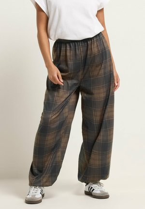 Persona che indossa pantaloni larghi a quadri marroni e neri con vita elastica, t-shirt bianca e sneakers bianche con strisce nere.