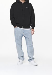 Felpa nera con zip e tasche frontali, abbinata a jeans azzurri larghi e sneakers bianche. La felpa presenta un logo sul petto.