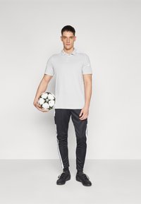 Svetlošedá polo tričko s gombíkovou légou, v kombinácii s čiernymi teplákmi so bielymi bočnými pruhmi a čiernymi športovými topánkami. Drží futbalovú loptu.