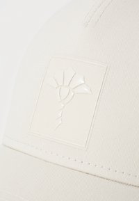 Hvid kasket med et struktureret design, der omfatter et tekstureret logo-applikation med bladdesign og synlige sømme på skyggen.