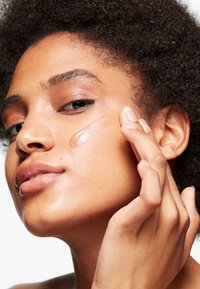 Fuktgivande serum applicerat på huden. Slät, glänsande textur. Närbild av ansikte med naturlig makeup, lockigt hår och väldefinierade drag.