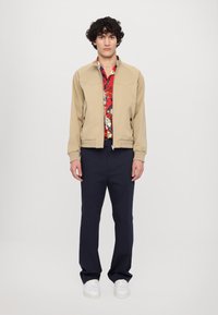 Beige lehká bunda na zip s vroubkovaným límcem a manžetami. Pod ní červená košile s květinovým vzorem a krátkými rukávy.