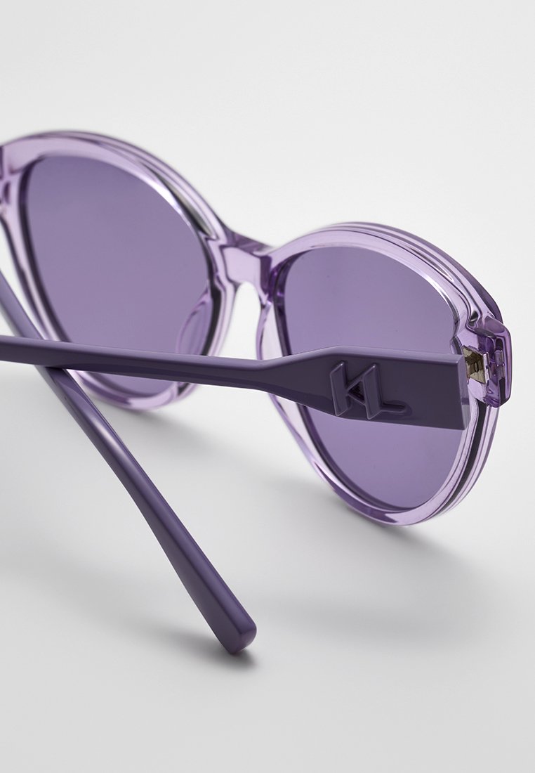 Lunettes de soleil violettes avec des verres ronds, monture transparente et un logo distinct sur la branche. Texture lisse et design moderne.