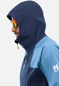 Dunkelblaue und hellblaue Outdoor-Jacke mit Kapuze, ausgestattet mit einem Reißverschluss und roten Akzenten; mit dem Markenlogo "MILLET" auf der Brust.