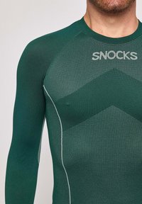 Haut de sport long à manches longues vert avec un motif texturé, présentant un col ras du cou et un logo réfléchissant. Des accents en vert plus clair offrent un contraste.