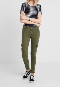 Pantalones cargo verde oliva con parches en las rodillas, corte slim, cinco bolsillos y dobladillos ajustados, combinados con una camiseta de rayas azul marino y blanca y zapatillas negras.