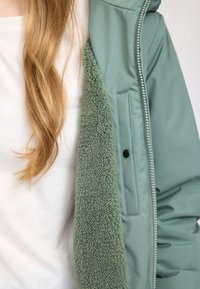 Grüne Zip-Jacke mit einer weichen Fleece-Fütterung. Hat eine glatte Außentextur, schwarze Beschläge und eine lockere Passform.
