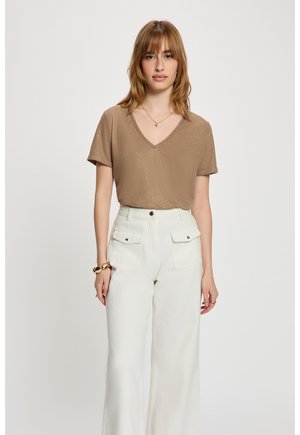 Mujer con cabello hasta los hombros que lleva un top marrón brillante con escote en V, pantalones blancos de cintura alta con bolsillos delanteros y joyas de oro en la muñeca y el cuello.