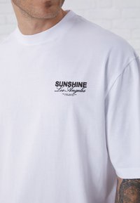 White cotton t-shirt with short sleeves, round neck, featuring a black printed logo "SUNSHINE Los Angeles" on the left chest.