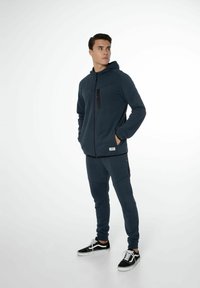 Haut à capuche zippé bleu marine avec poche zippée noire, associé à un pantalon de jogging assorti. Le tissu semble doux avec des détails en côte aux poignets et à l'ourlet.