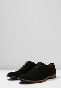 Chaussures en daim noires à lacets avec un bout rond, une tige texturée et une semelle brune. Elles présentent des lacets fins ton sur ton et un intérieur lisse.