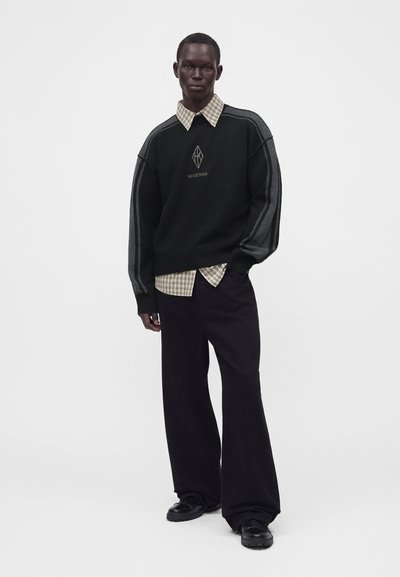 Homme portant un pull noir avec des rayures grises et un logo par-dessus une chemise beige à carreaux, un pantalon large noir et des chaussures noires, debout avec une main dans la poche.
