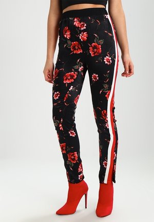 Le modèle porte des leggings noirs à fleurs avec des bandes latérales rouges et blanches ainsi que des bottines rouges vives à talons hauts.