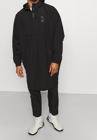 Schwarzer leichter Anorak mit einem Halbreißverschluss, verstellbare Kapuze und Reißverschlusstaschen vorne. Kombiniert mit schwarzen Jogginghosen und weißen Sportschuhen.