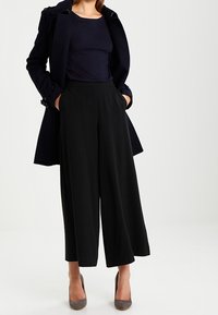 Pantaloni neri a vita alta e a gamba larga con tasche laterali, abbinati a un top aderente blu navale e a un lungo cappotto scuro, completati da scarpe con tacco grigie.