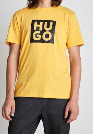 Osoba nosząca żółtą koszulkę z krótkim rękawem z czarnym kwadratowym logo „HUGO” na piersi, w połączeniu z ciemnymi spodniami.
